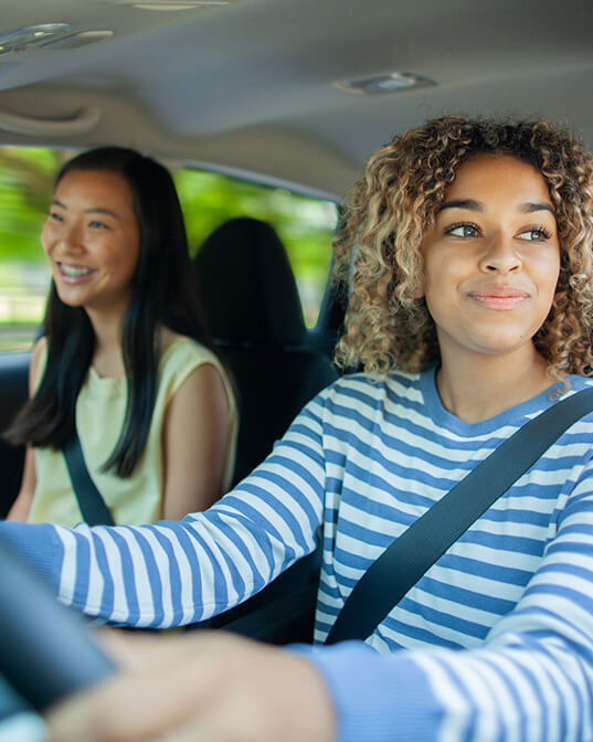 Driver grips the steering wheel, smiling, while a passenger sits beside her. Both wear seatbelts inside a moving car. Sunlight filters through the window, creating a cheerful atmosphere.