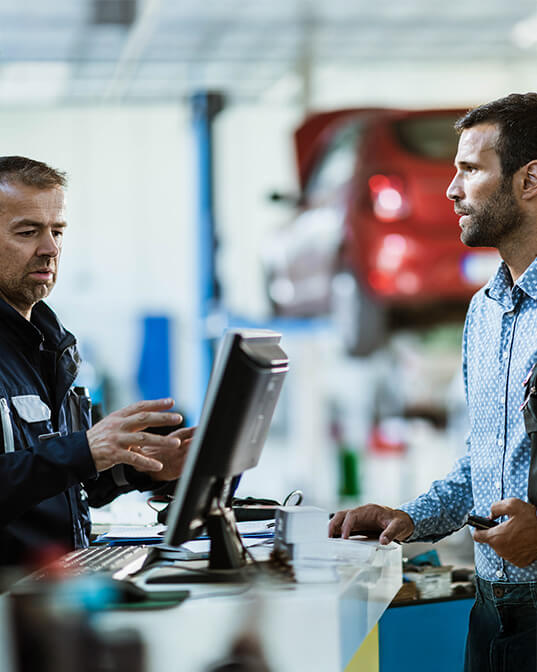 Two men converse at a service desk; one gestures while speaking to the other, who listens intently. In the background, a car is raised on a lift.