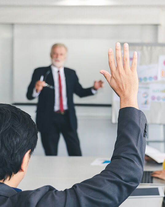 A person raises their hand in the foreground, while a man in a suit gestures during a presentation, standing in front of a whiteboard in a classroom or meeting room setting.